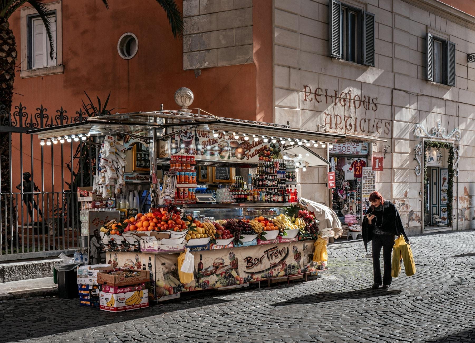 A street food and market stall in Rome selling fruit, snacks, and drinks, set against a classic Roman building with people passing by.