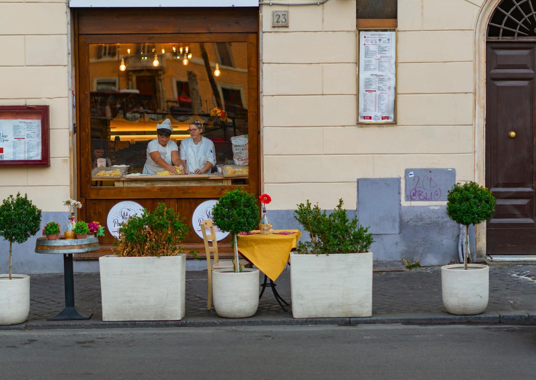 Street view of a Roman trattoria with chefs visible through the window preparing food, potted plants outside, reflecting authentic local dining.