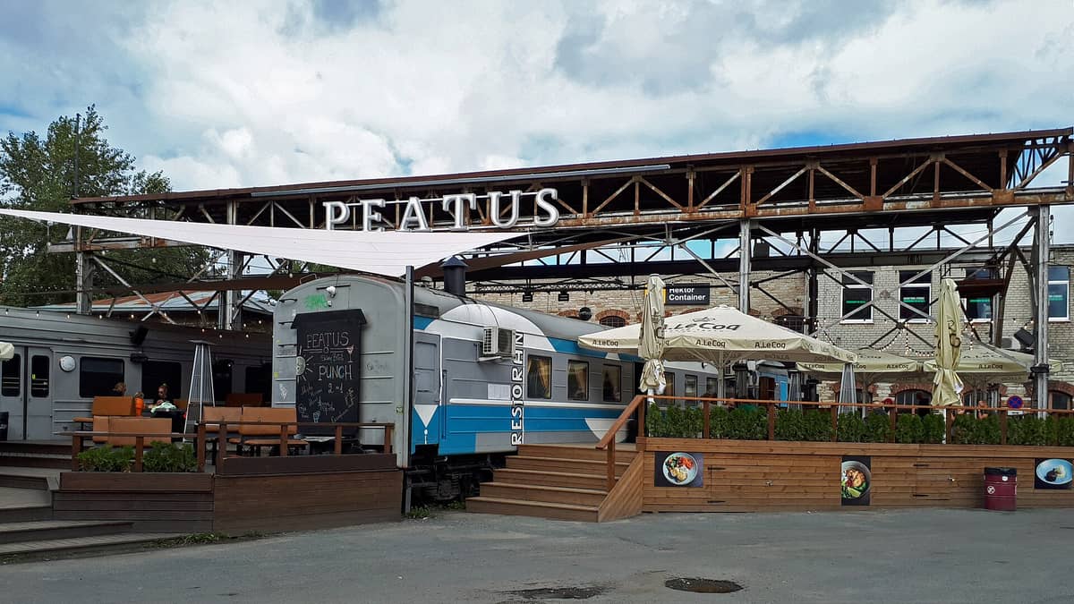 Außenansicht des Restaurants Peatus in Telliskivi, Tallinn, mit Eisenbahnwaggons als Speisewagen und Industriestrukturen mit Personen an Tischen