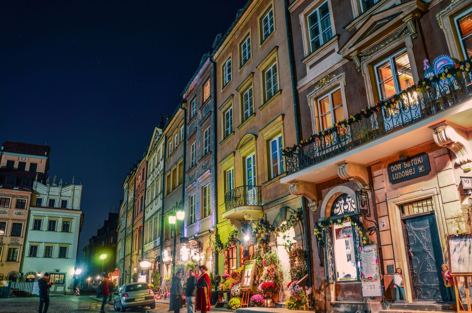 Vista noturna de uma rua animada de Varsóvia com coloridos edifícios históricos e restaurantes enfeitados com flores, capturando a vibrante atmosfera gastronômica da cidade.