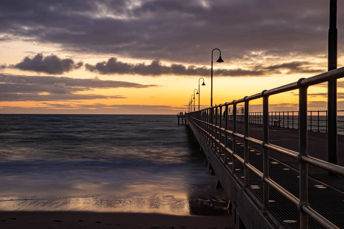 Langer Steg, der sich bei Sonnenuntergang ins Meer erstreckt, mit Sandstrand im Vordergrund und sanften Wellen unter einem bewölkten Himmel.