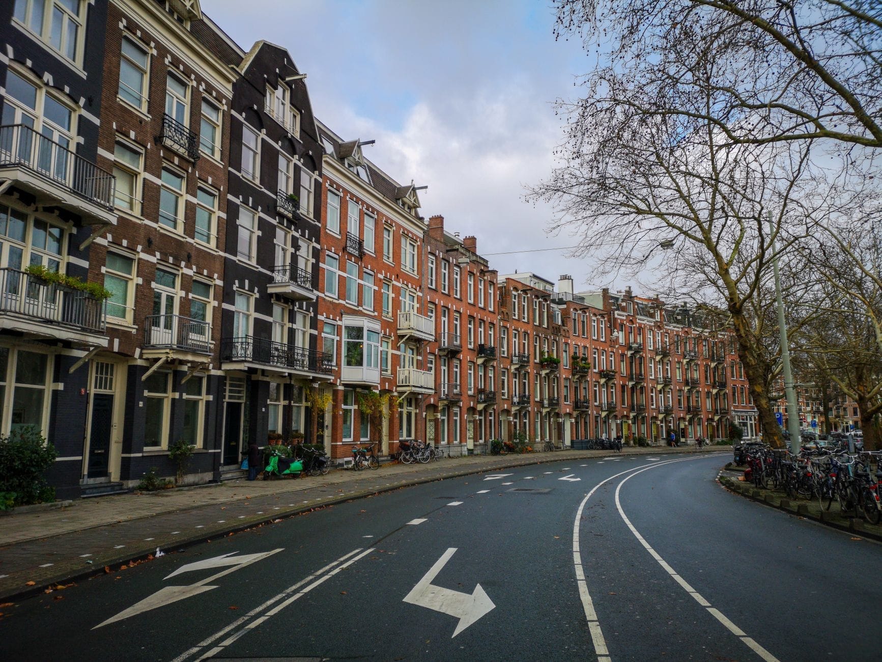 Amplia calle de Ámsterdam bordeada de casas adosadas de ladrillo tradicionales, árboles y bicicletas aparcadas a lo largo de la acera en un día nublado.