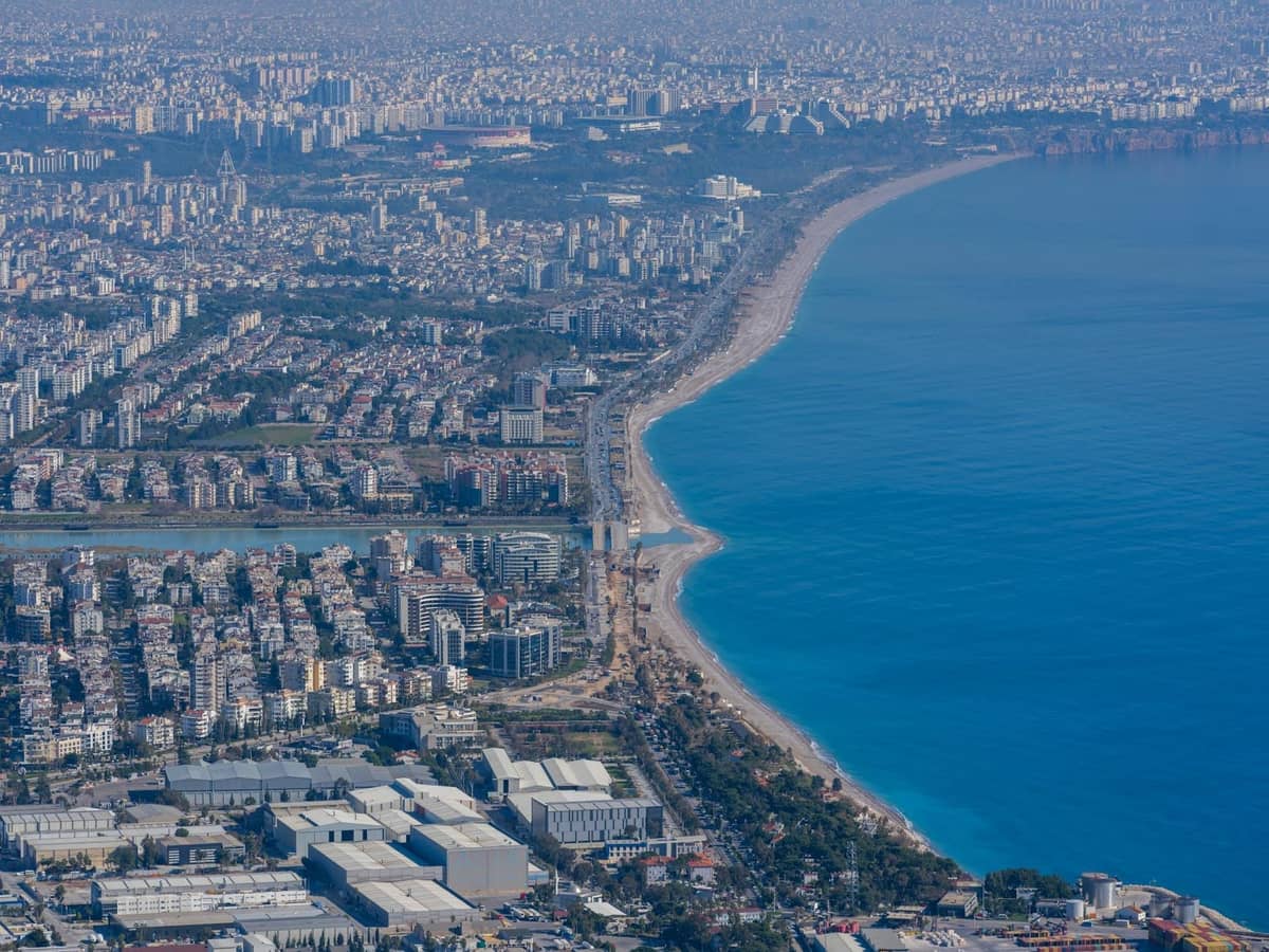 Aerial view of Antalya showing long curving coastline, dense cityscape, and beach along the Mediterranean Sea.