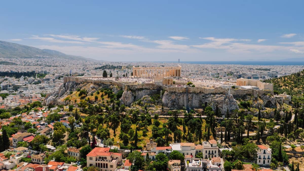 Pemandangan udara Acropolis dan kawasan sekitarnya, termasuk area hijau, bangunan perumahan, dan lanskap kota di bawah langit biru.