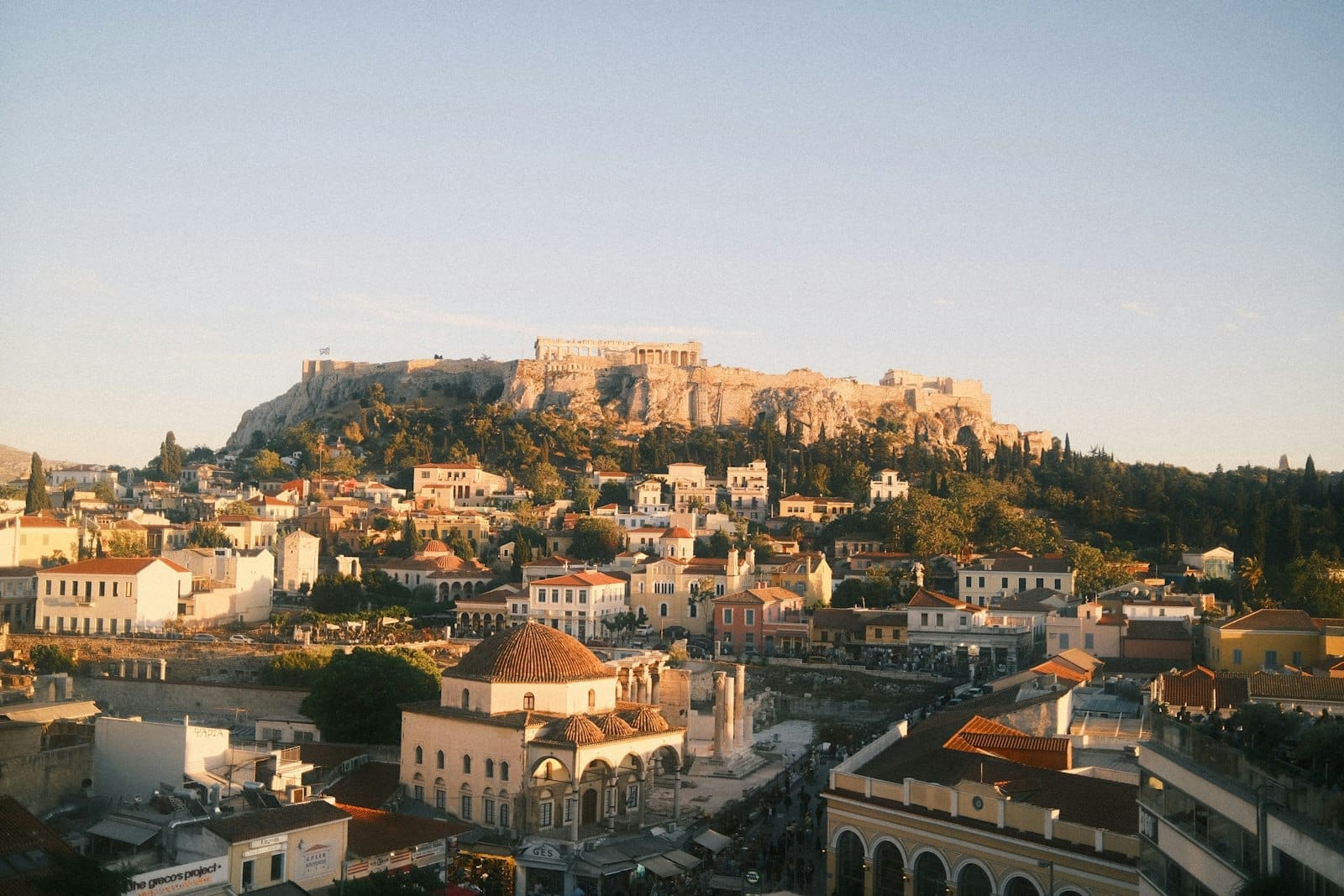 Vista ampla do centro de Atenas com a Acrópole emergindo acima dos telhados da cidade na hora dourada, capturando bairros urbanos e pontos icônicos sob um céu limpo.