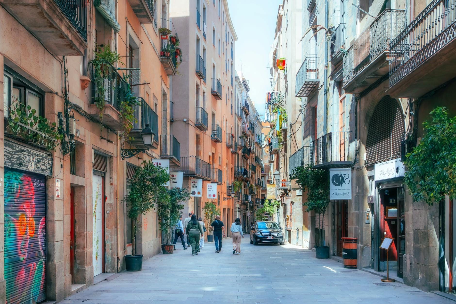 Colorful, lively pedestrian street in El Born Barcelona with locals, balconies, potted plants, and characteristic buildings under bright daylight.
