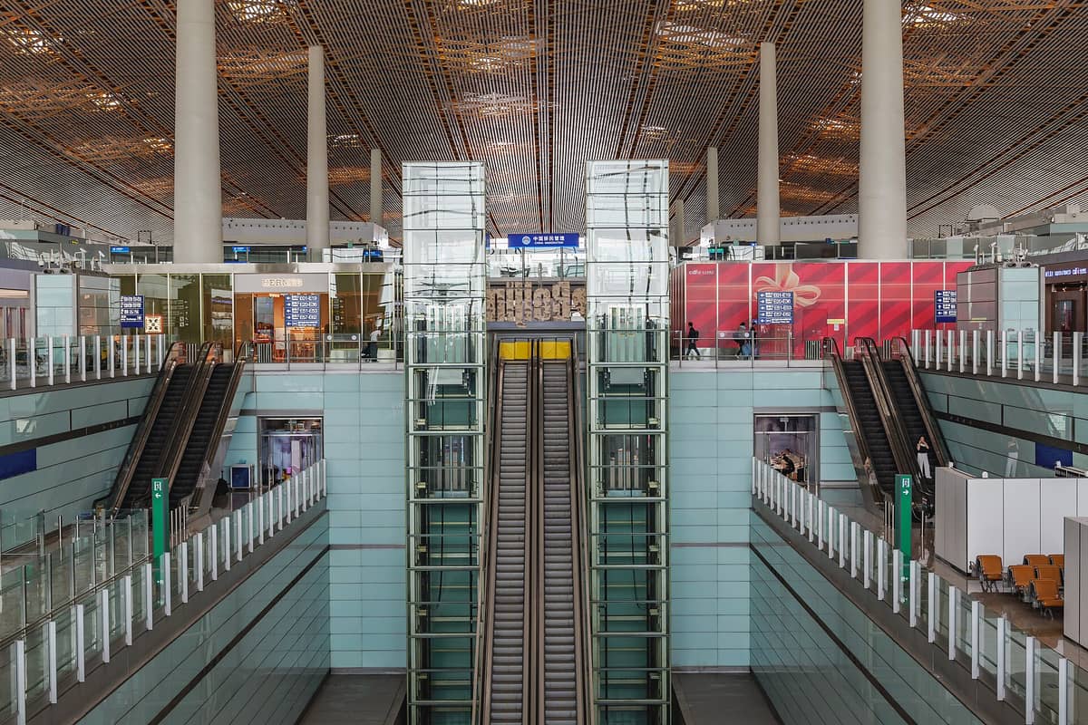 Interior of Beijing Capital International Airport terminal with escalators, glass elevators, and signage under a modern patterned ceiling.
