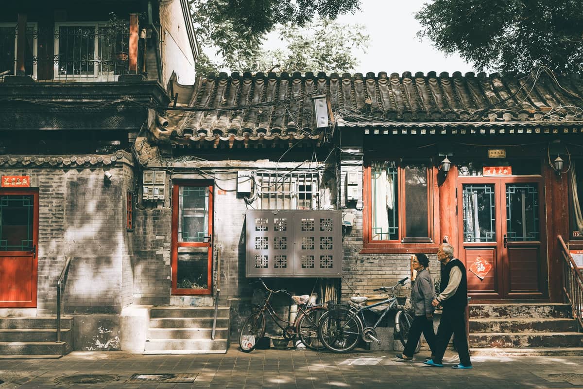 Two people walk past traditional hutong-style buildings with tiled roofs, red doors, and bicycles in leafy shade in Beijing.