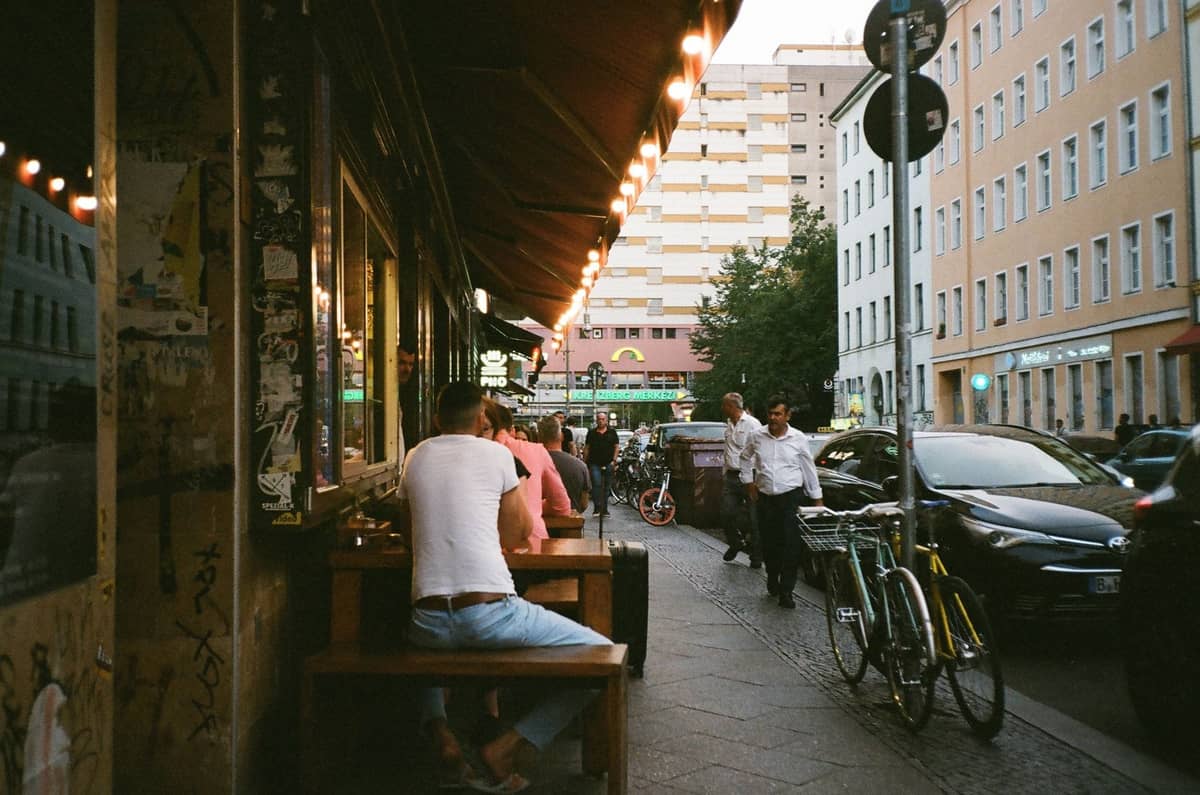Scène animée sur un trottoir avec des personnes assises à des tables en terrasse, des vélos, des immeubles d'appartements et l'agitation de la rue, typique des quartiers branchés de Berlin.