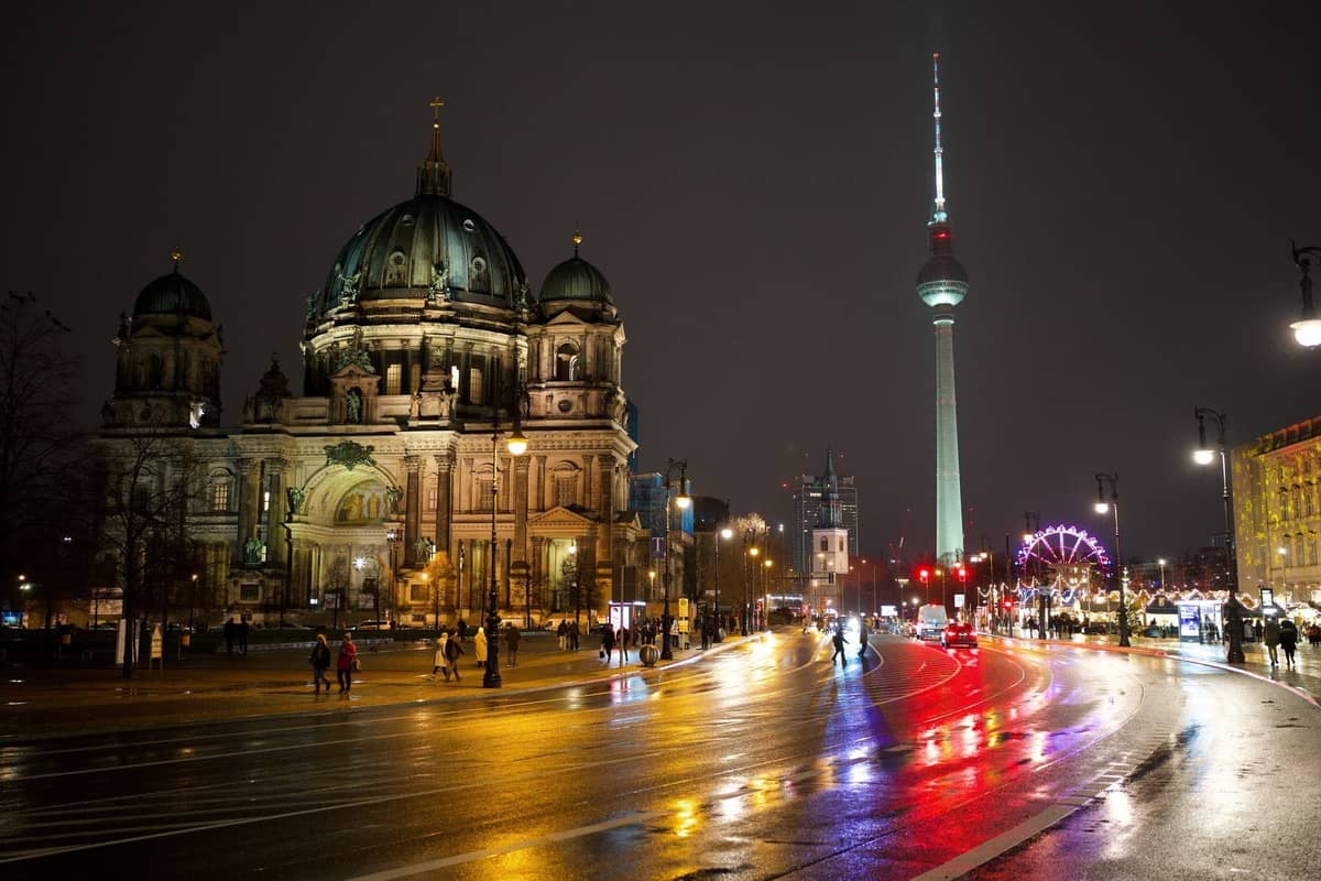 Vue nocturne de la Cathédrale de Berlin et de la Fernsehturm, avec des lumières se reflétant sur les rues mouillées et des personnes se promenant à proximité.