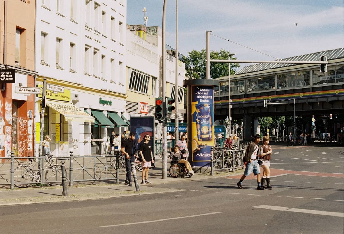 Scène de rue dans un quartier de Berlin avec des personnes qui marchent, des cafés en plein air, des graffitis et des voies de train surélevées, capturant une atmosphère urbaine animée.