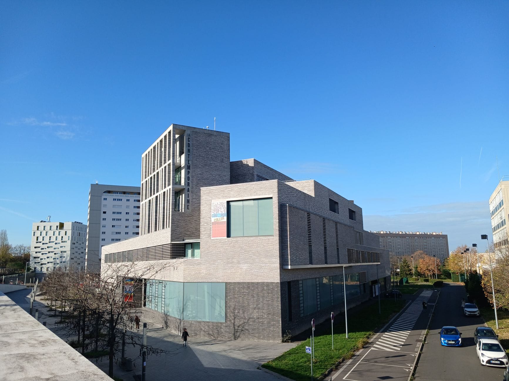 Modern building in a quiet residential area with nearby roads and a few cars, under a clear blue sky.