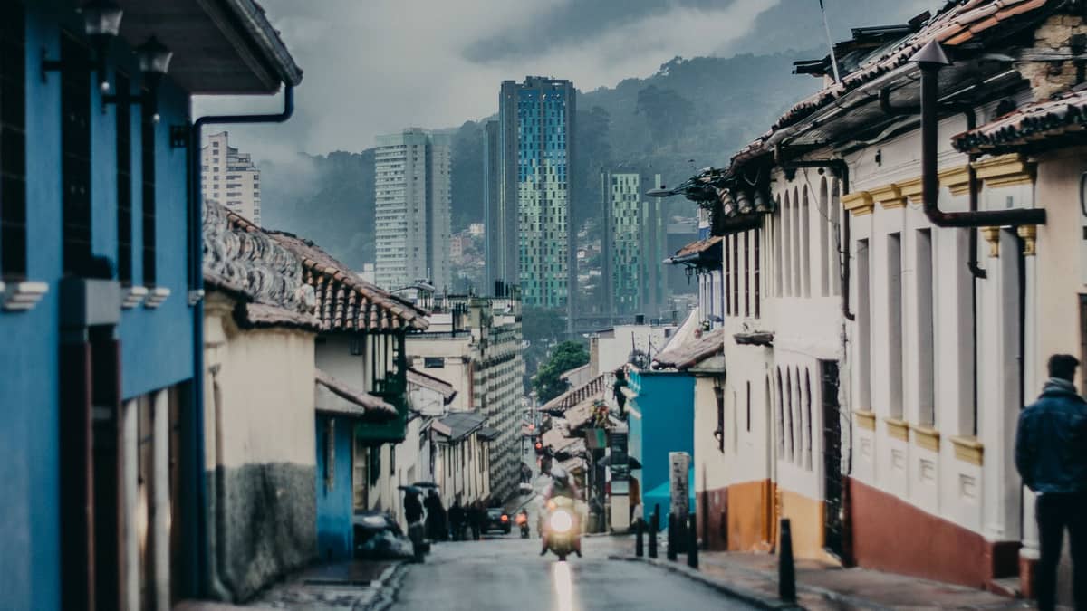 Colorful colonial street in La Candelaria, Bogotá, with historical houses in foreground and modern buildings in the background.