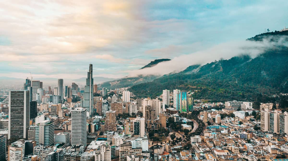 A wide aerial view of Bogotá’s skyline with tall buildings against green mountains and dramatic clouds