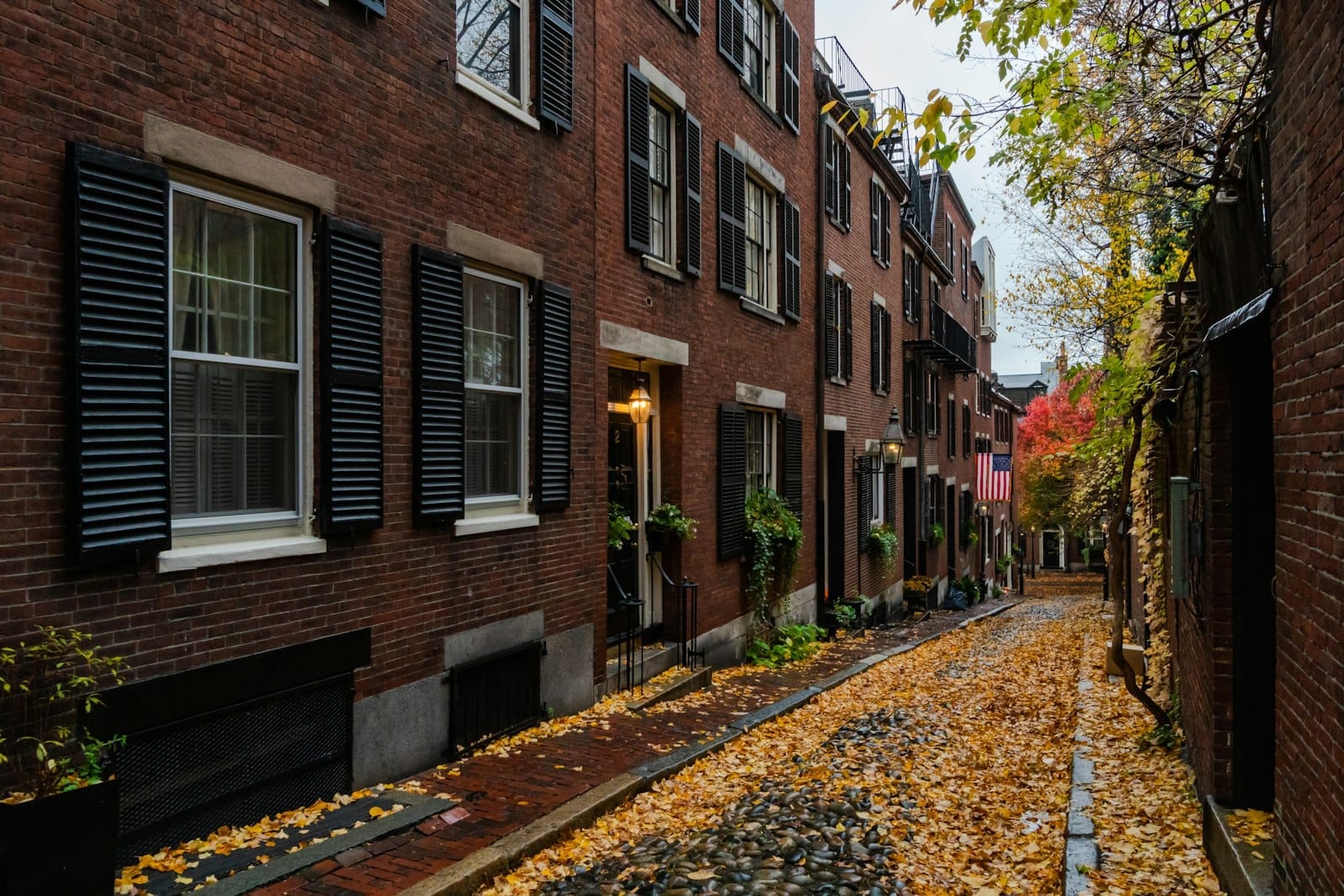 A histórica Acorn Street no bairro Beacon Hill de Boston, com casas geminadas de tijolo, venezianas pretas, calçamento de paralelepípedos e folhas de outono espalhadas pela calçada.