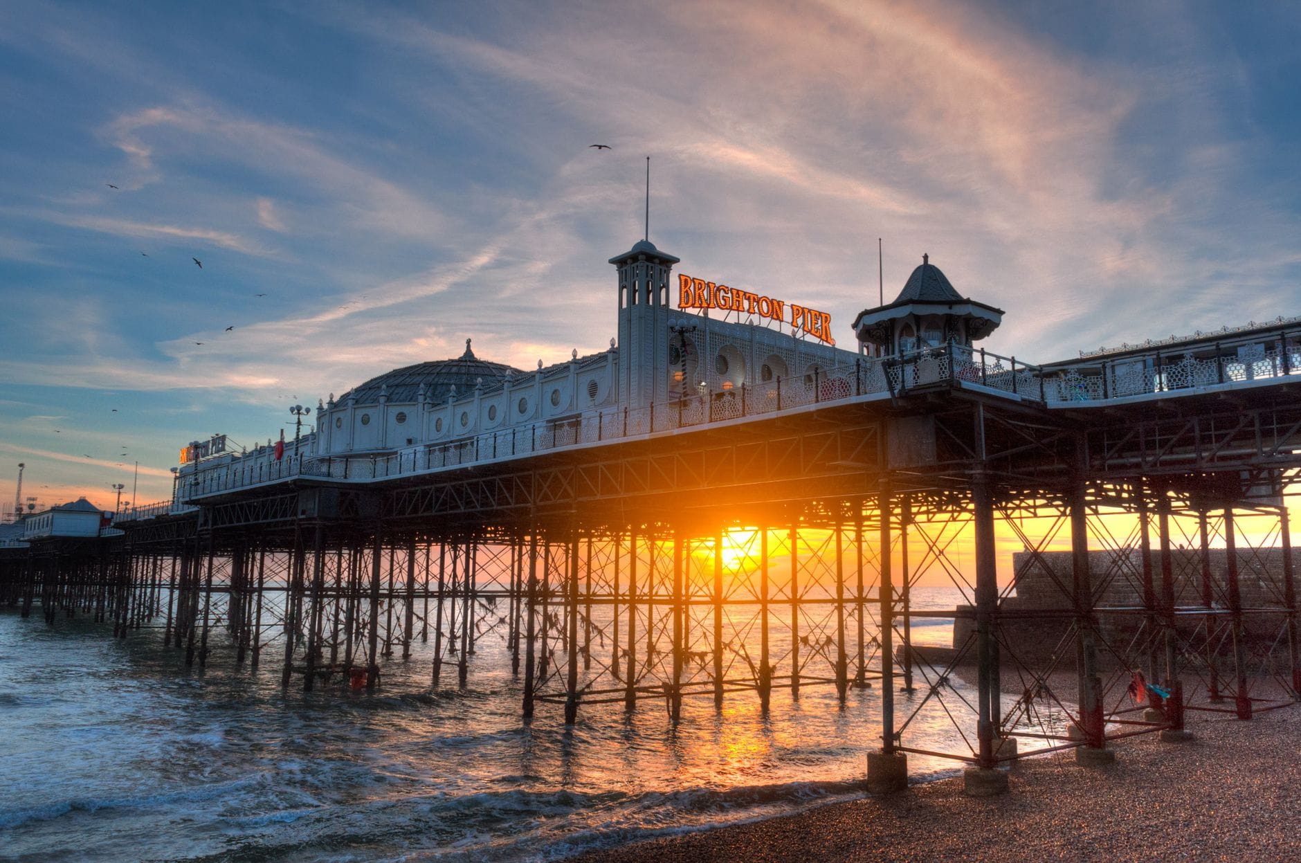 Brighton Pier at sunset with the sun glowing through the pier structure, waves gently hitting the shore, and a colorful sky overhead.