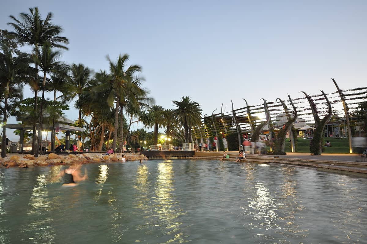 Landscape view of South Bank Parklands' lagoon pool at dusk, with palm trees, stone-edged water, and people relaxing near the water in Brisbane.