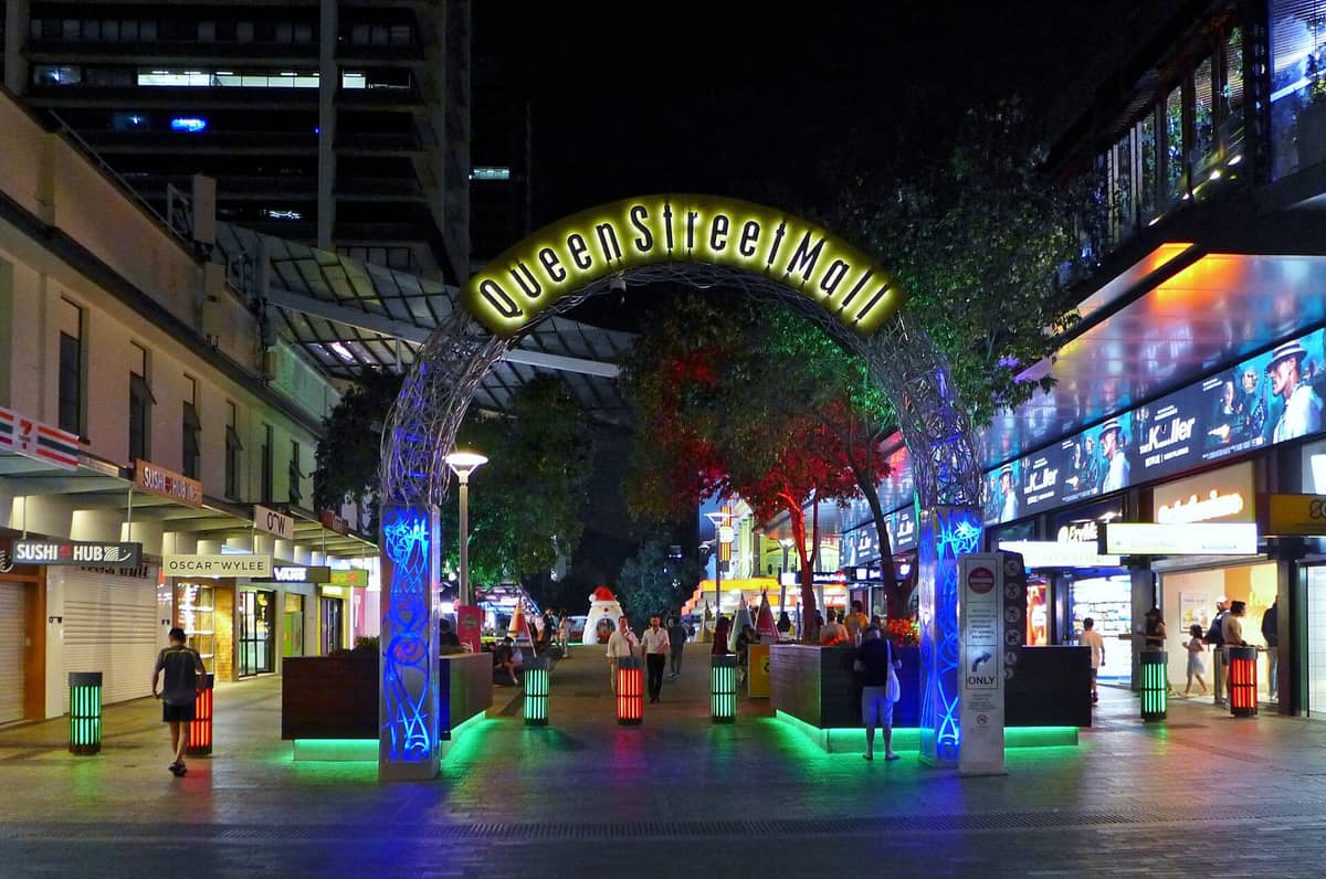 Night view of Queen Street Mall entrance in Brisbane CBD, brightly lit with neon lights, shops, and people walking around.