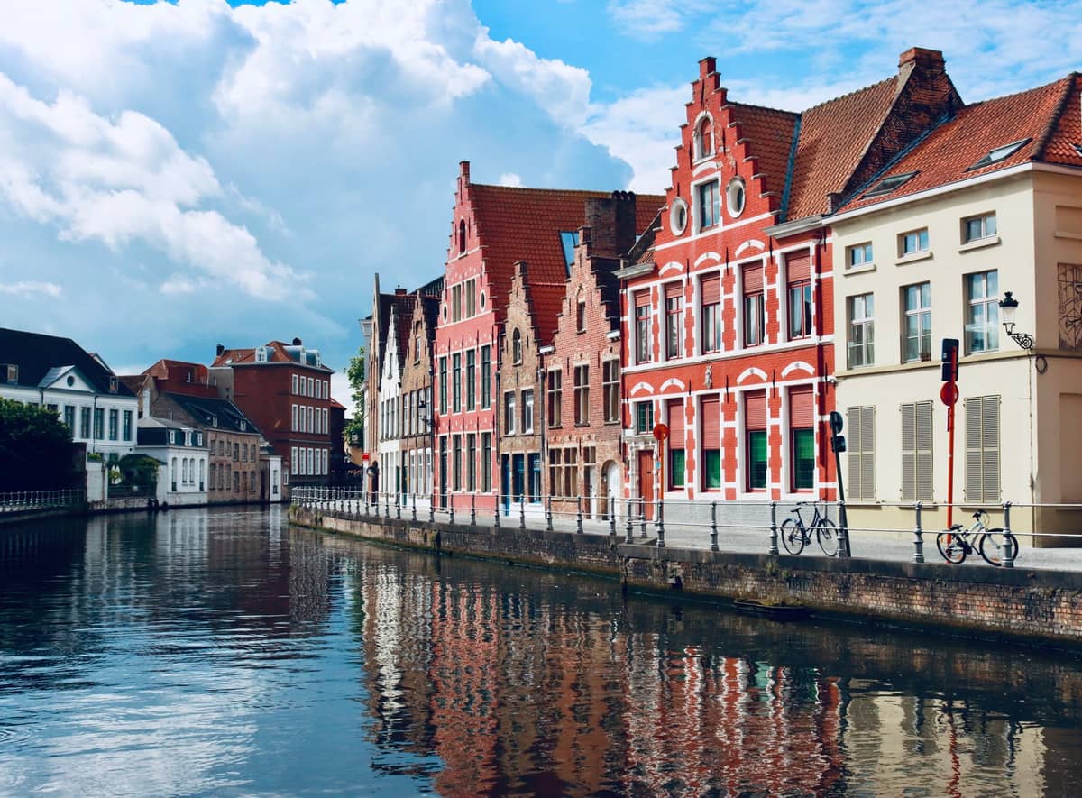 Row of colorful traditional riverside houses in Bruges, Belgium, with bicycles parked along the canal, under a bright sky.