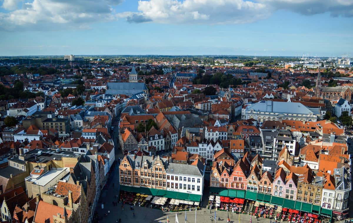 A wide aerial view of Bruges with iconic stepped-gable buildings and medieval rooftops, displaying neighbourhoods in the city’s historic center under a partly cloudy sky.