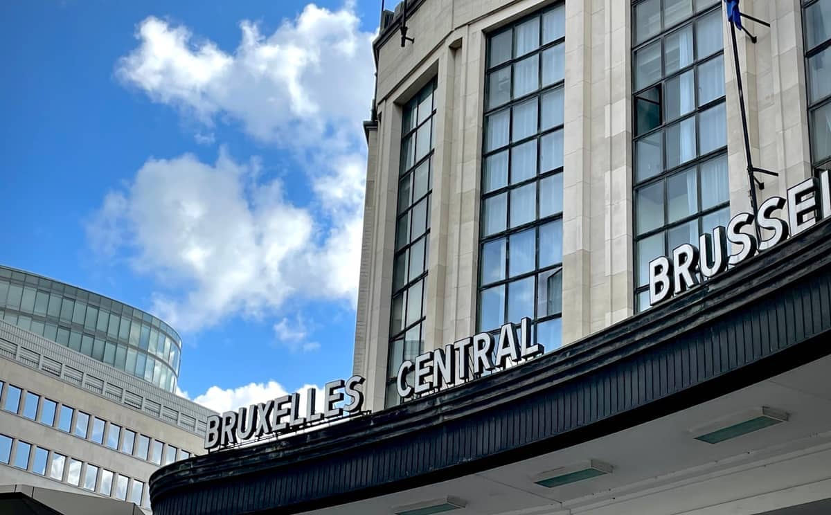 Vista diurna da fachada da estação de comboios Bruxelles Central com arquitetura moderna, grandes janelas e céu azul brilhante com nuvens esparsas.
