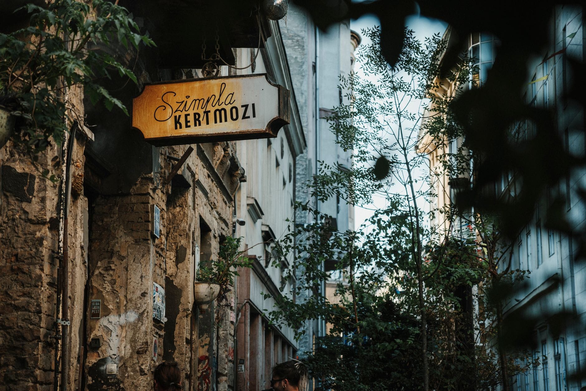 Szimpla Kertmozi-Schild in einer Ruinenkneipengasse, umgeben von Laub und alten Gebäuden im jüdischen Viertel von Budapest.