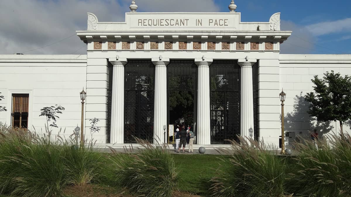 ทางเข้าหลักของสุสาน Recoleta ที่มีเสาสีขาว ประตูเหล็กลวดลายประณีต และจารึก 'REQUIESCANT IN PACE' ล้อมรอบด้วยต้นไม้สีเขียวและท้องฟ้าสีฟ้า