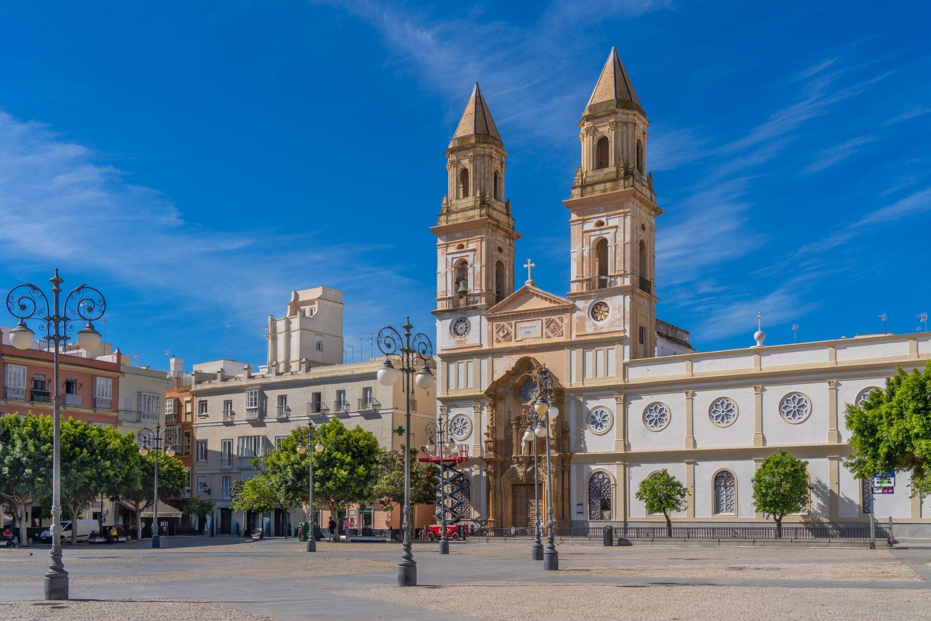 Wide plaza with ornate lampposts, trees, colorful historic buildings, and twin-towered church in Cádiz old town under a clear blue sky.