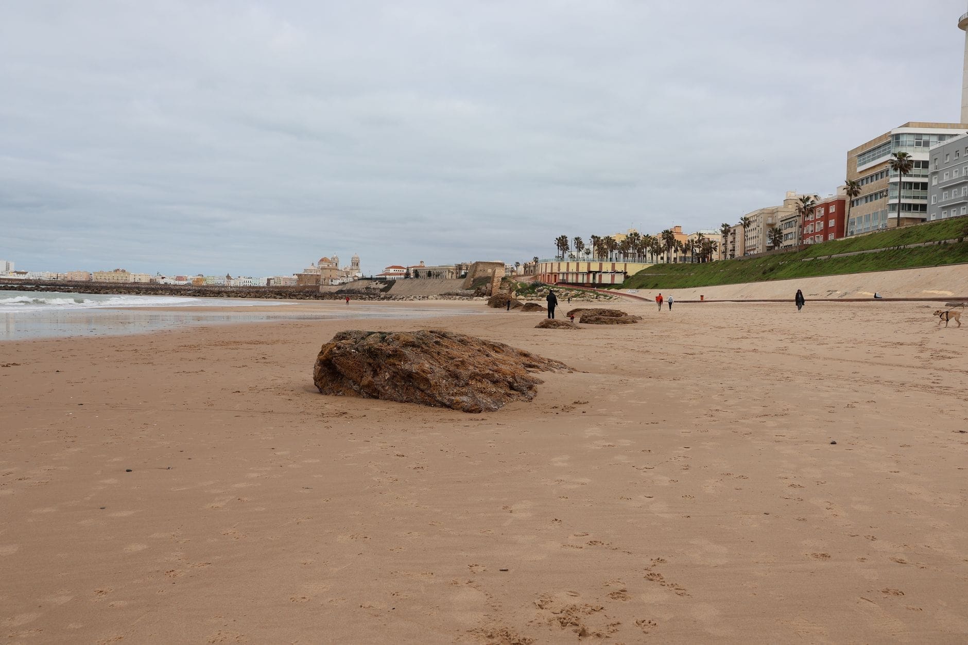 Wide sandy beach with scattered rocks, people walking, and modern seafront hotels lining the promenade in Cádiz, Spain.