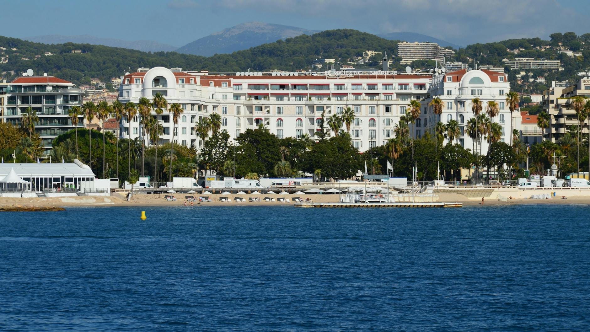 Grande hotel de frente para a praia de Cannes com palmeiras, espreguiçadeiras e o mar azul ao fundo sob um céu ensolarado.