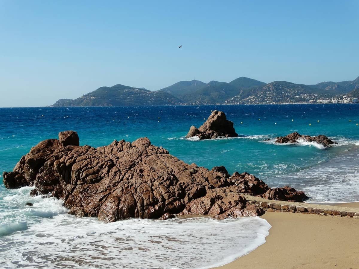 ชายหาดทรายเงียบสงบที่ Cannes La Bocca พร้อมโขดหินในเบื้องหน้า ทะเลสีฟ้า และเนินเขาที่อยู่ไกลออกไปใต้ท้องฟ้าแจ่มใส
