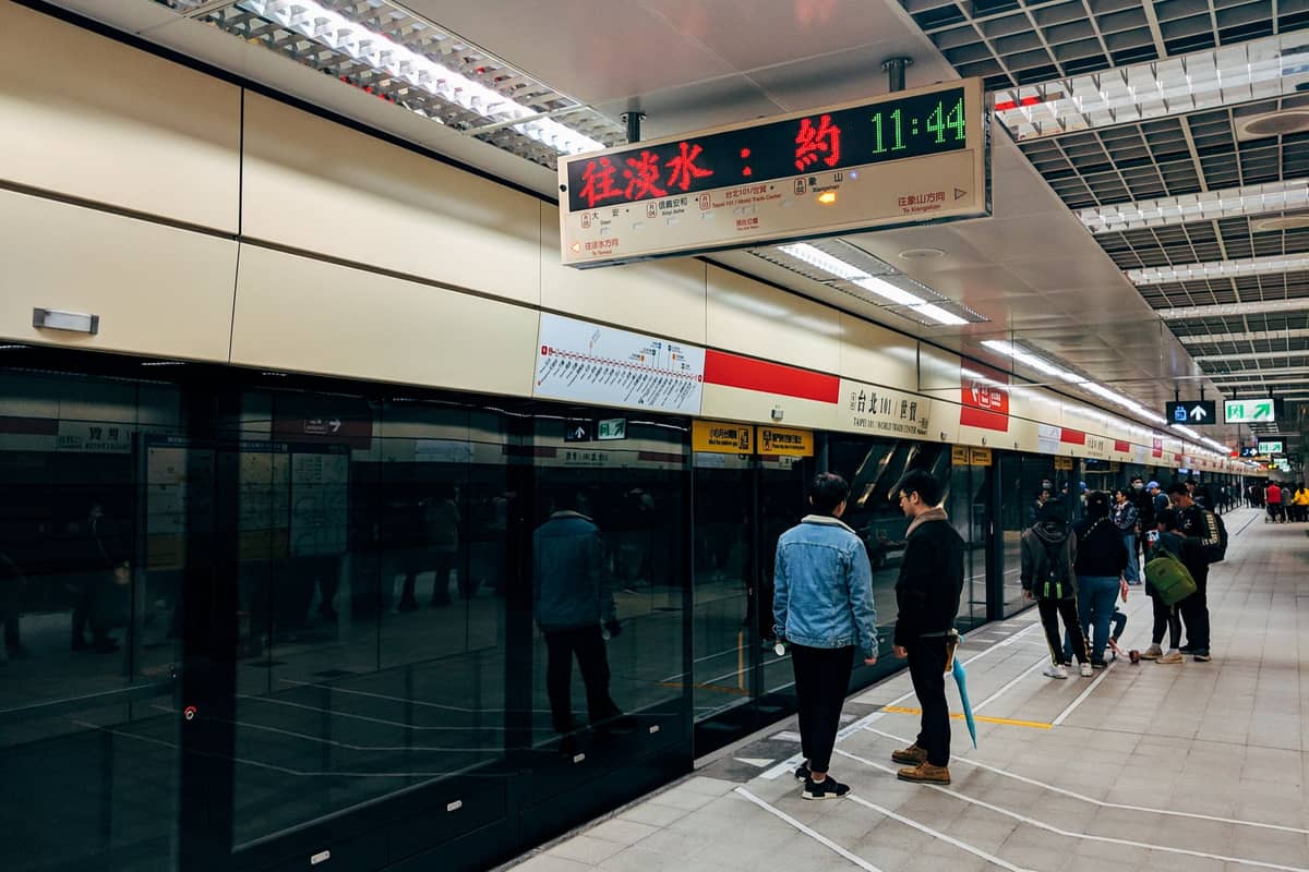Люди ожидают на современной платформе китайского метро со стеклянными барьерами и электронными табло над головой.
