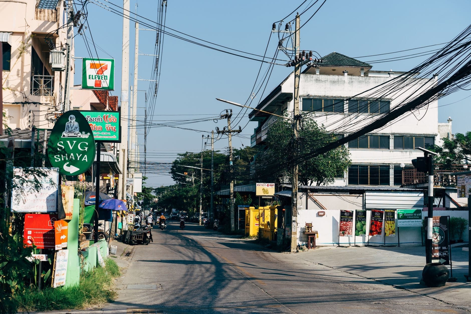 Una calle urbana en Chiang Mai bordeada de pequeñas tiendas locales, cafés, un 7-Eleven, letreros de restaurantes y cables eléctricos visibles.