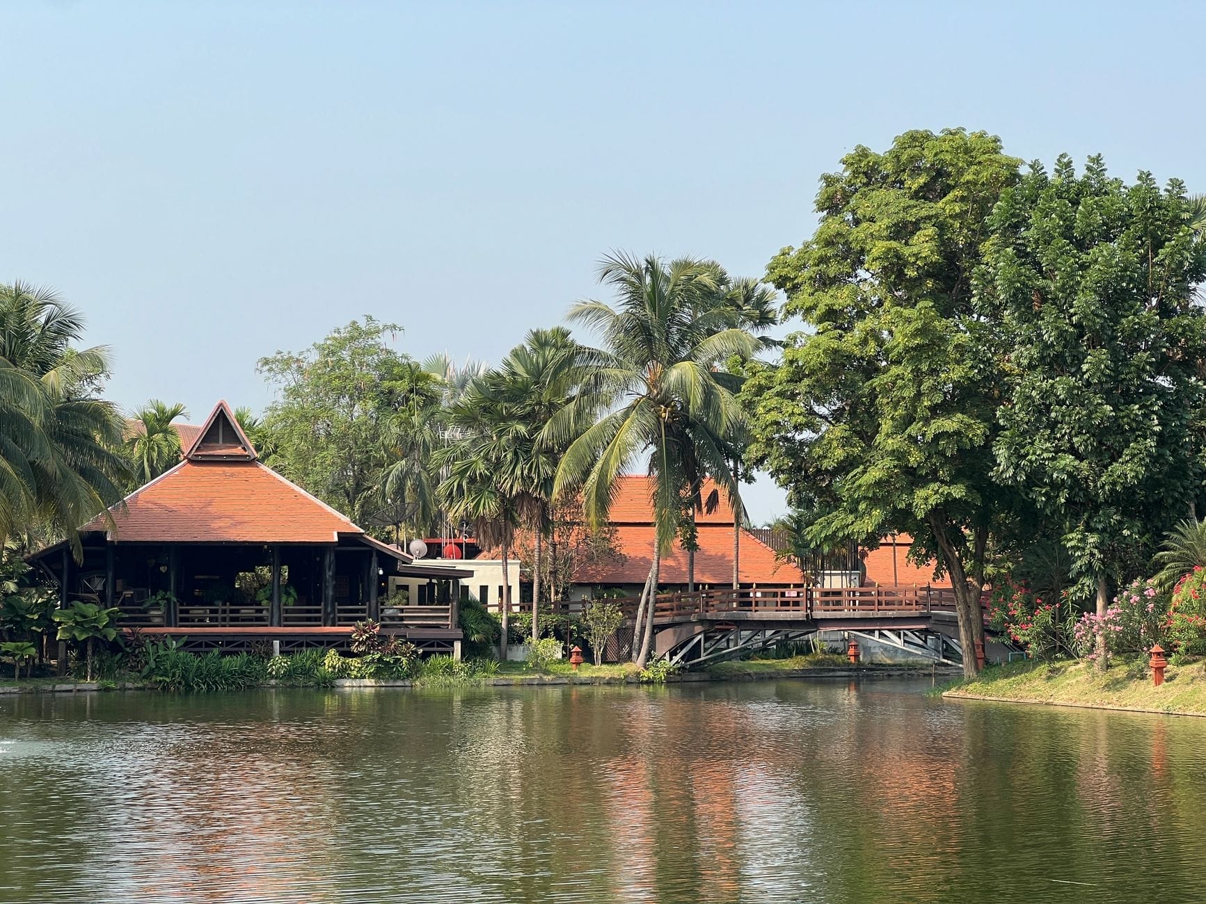 Edificios tradicionales de estilo tailandés a orillas del río con tejados rojos y exuberantes palmeras reflejadas en el agua tranquila en un día soleado.