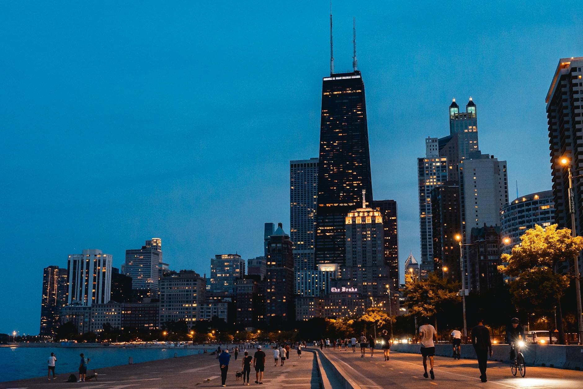 Vista noturna do skyline de Chicago com prédios iluminados, pessoas caminhando pelo calçadão à beira do lago e uma atmosfera urbana vibrante e acolhedora.