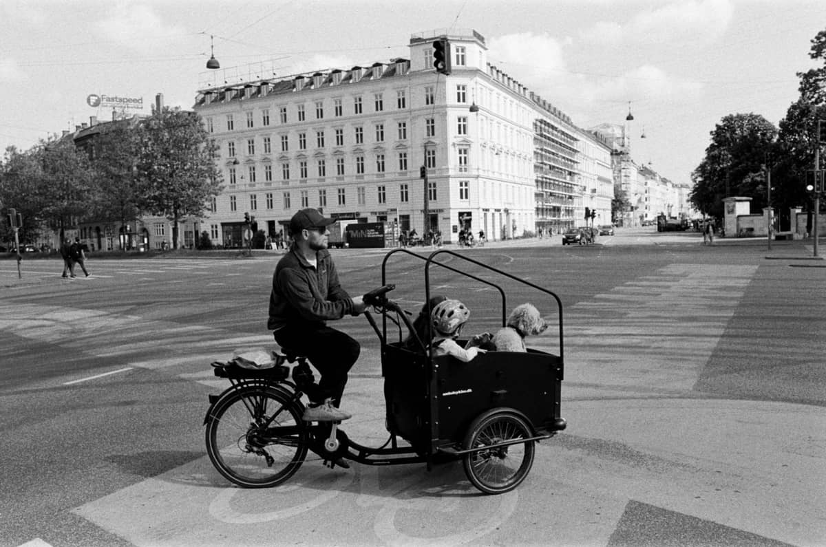 Schwarz-Weiß-Foto einer Person, die mit zwei Kindern in einem Lastenrad eine städtische Kreuzung mit historischen Gebäuden überquert.