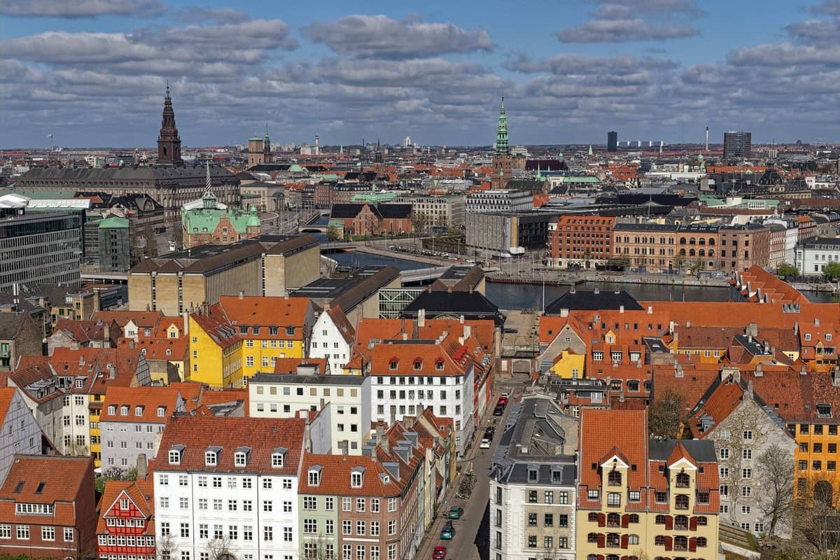 Ein weiter Luftblick über Kopenhagen mit markanten Vierteln, historischen Gebäuden, bunten Fassaden und zentralen Wahrzeichen unter einem teils bewölkten Himmel.