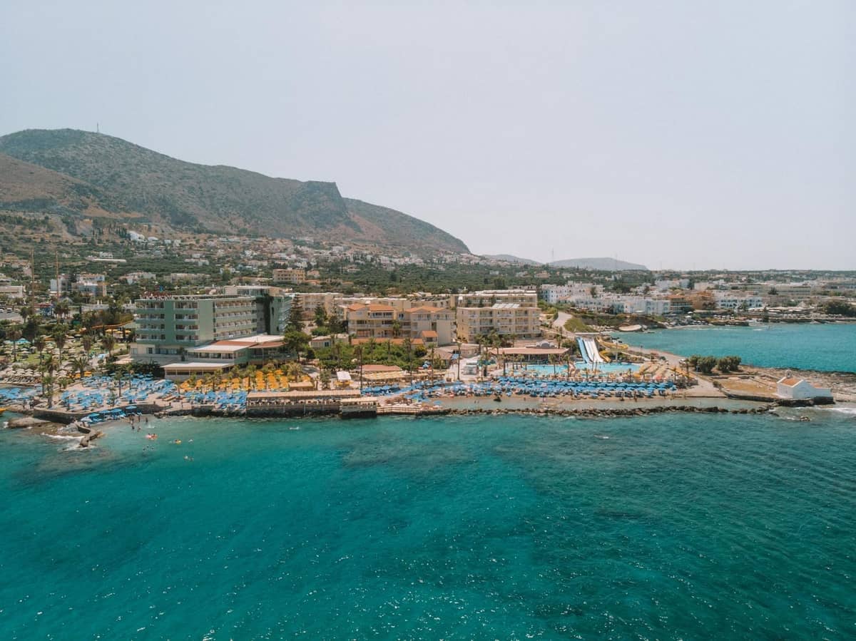 Aerial na tanawin ng baybayin ng Hersonissos na may malalaking hotel, sun lounger, pool, at makulay na waterfront na katangian ng resort strip ng Crete.