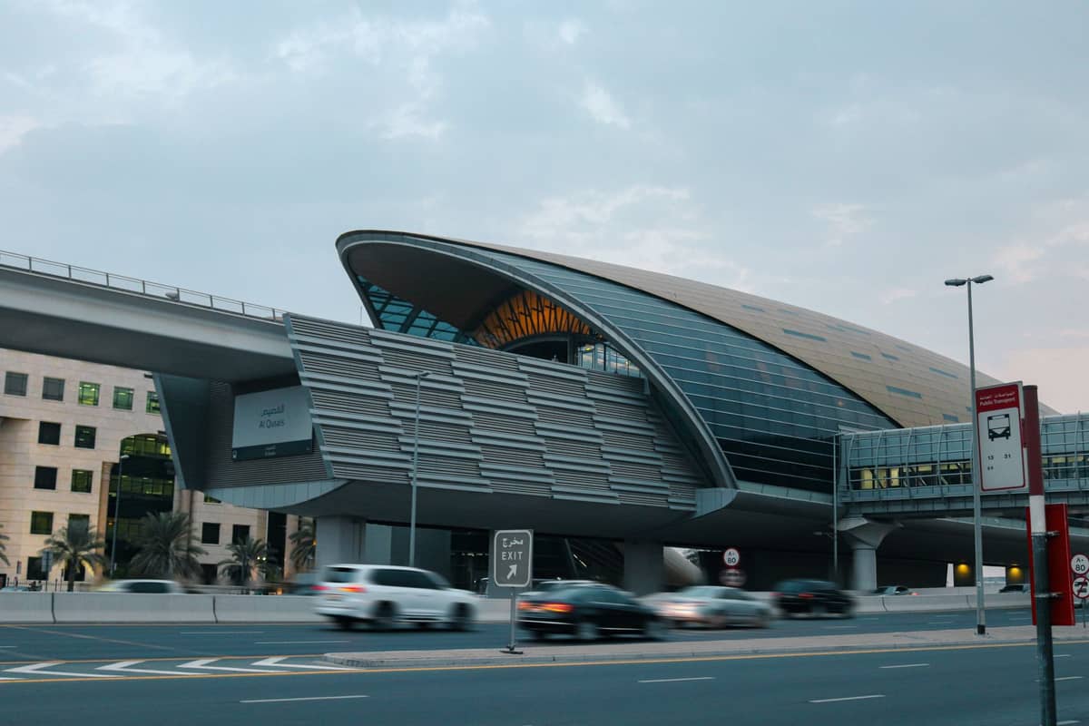 ภายนอกที่ทันสมัยของสถานี Dubai Metro พร้อมรถยนต์แล่นผ่านด้านหน้าและมองเห็นส่วนหนึ่งของรางยกระดับ
