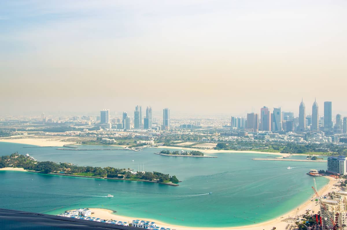 มุมมองกว้างของแนวชายฝั่ง Dubai ที่แสดงให้เห็นตึกระฟ้าสมัยใหม่ Palm Jumeirah และย่านริมน้ำหลายแห่งภายใต้ท้องฟ้าที่มีหมอกควัน
