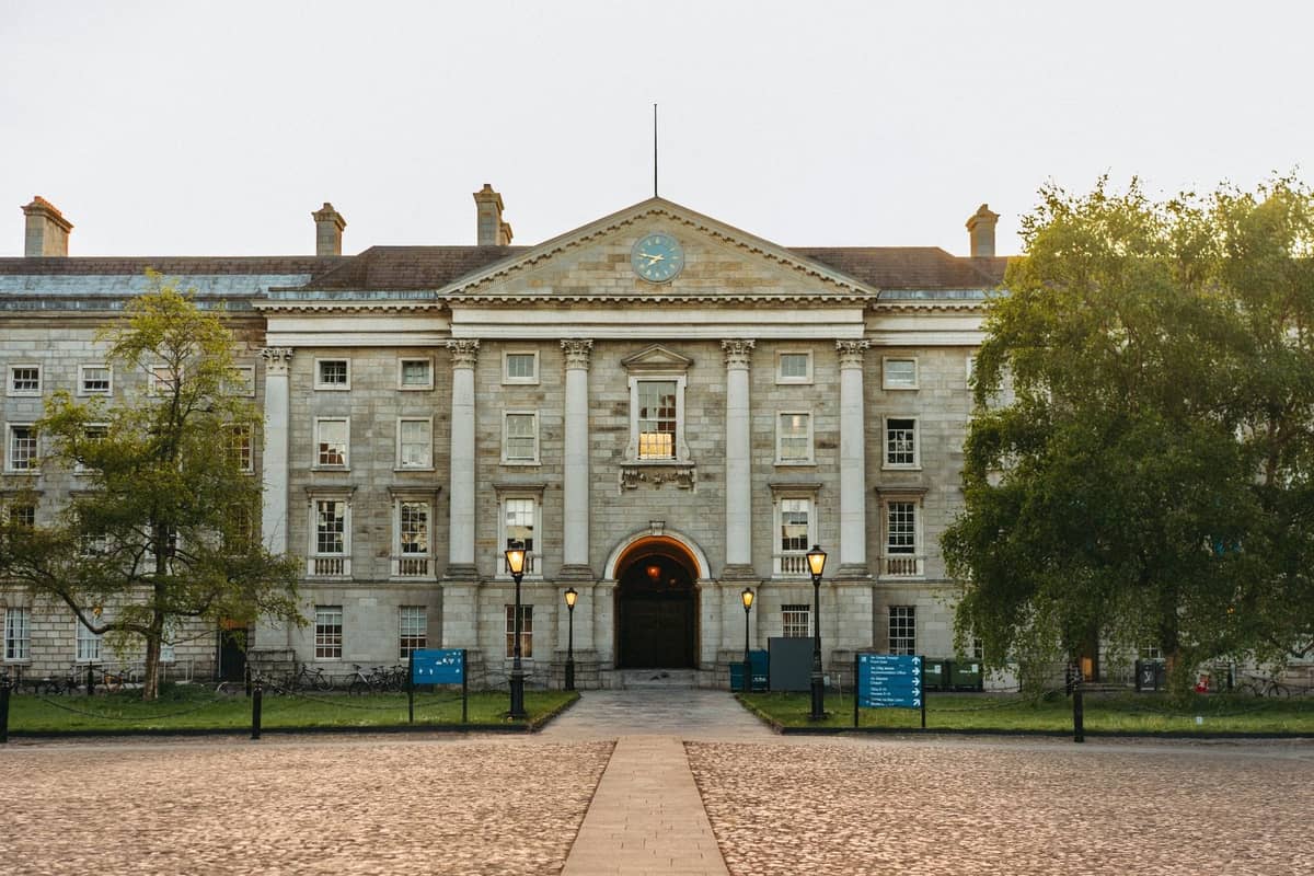 Front view of the historic stone facade of Trinity College Dublin with lamp posts and trees in the foreground.