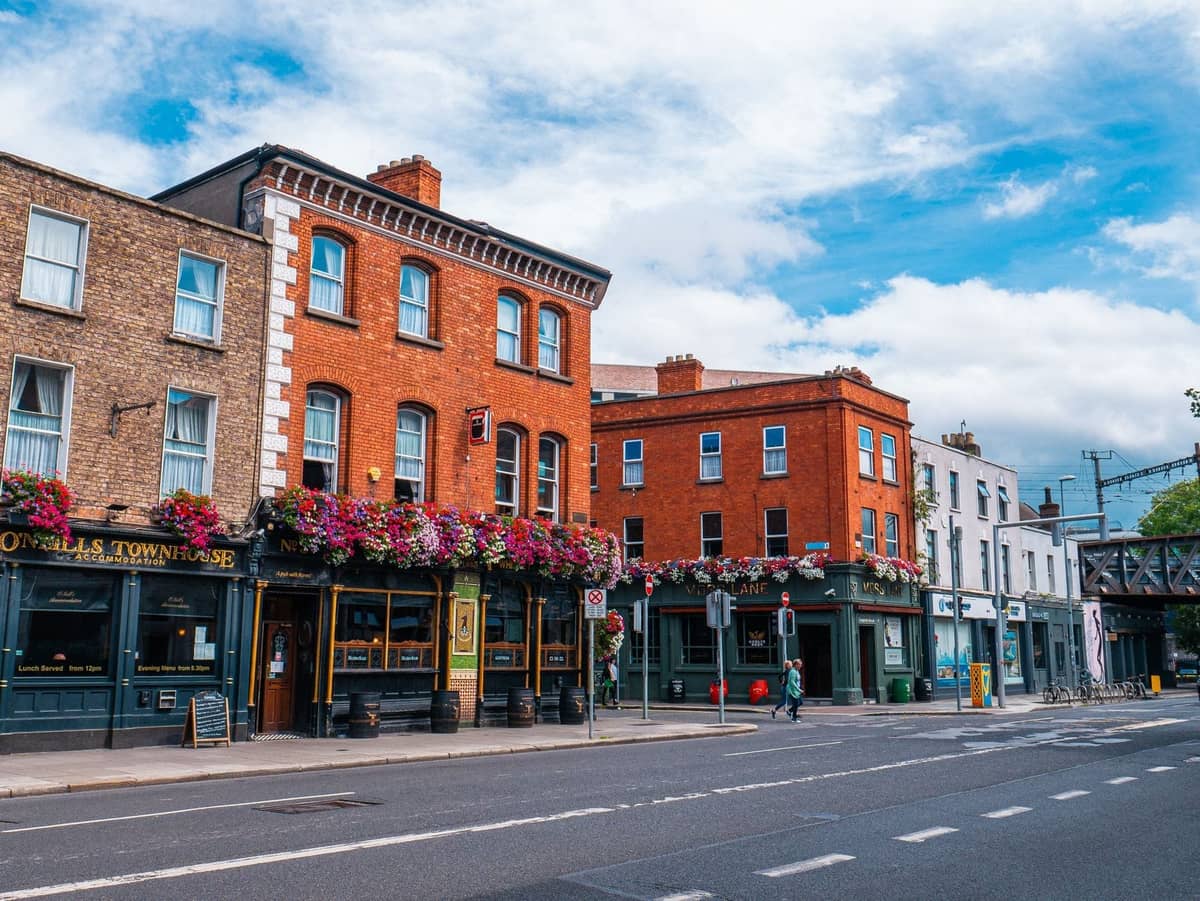 Street view of red-brick buildings, colorful flower baskets, and a pub in a leafy Dublin neighborhood with light traffic.