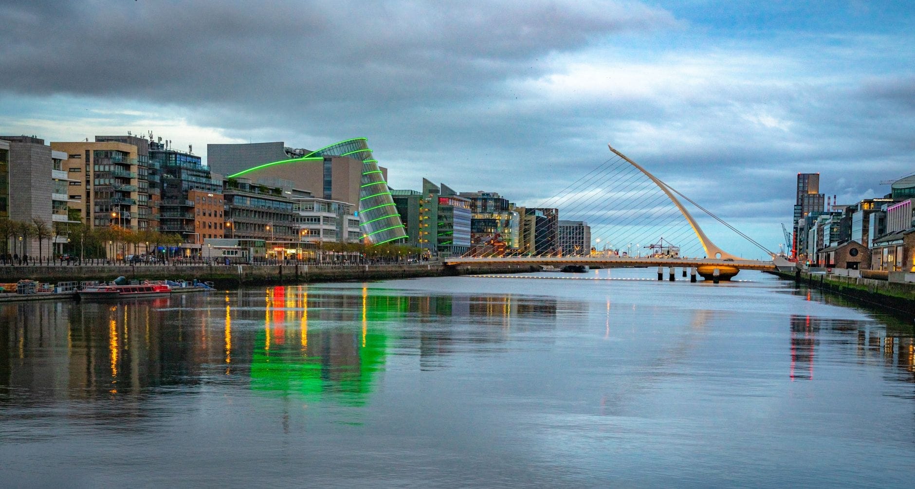 Vista ampla do horizonte de Dublin ao longo do Rio Liffey com a Ponte Samuel Beckett e prédios modernos sob um céu claro ao entardecer.