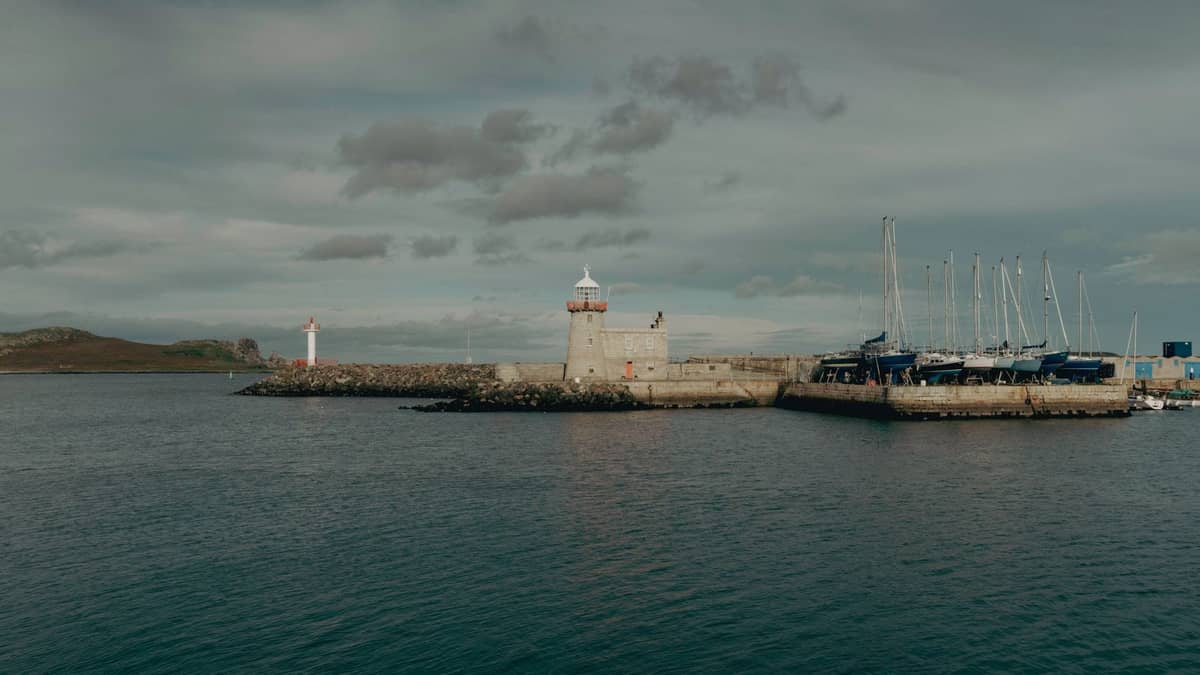 A wide view of Howth Harbour with its iconic lighthouse, stone piers, and moored sailboats on a cloudy day.