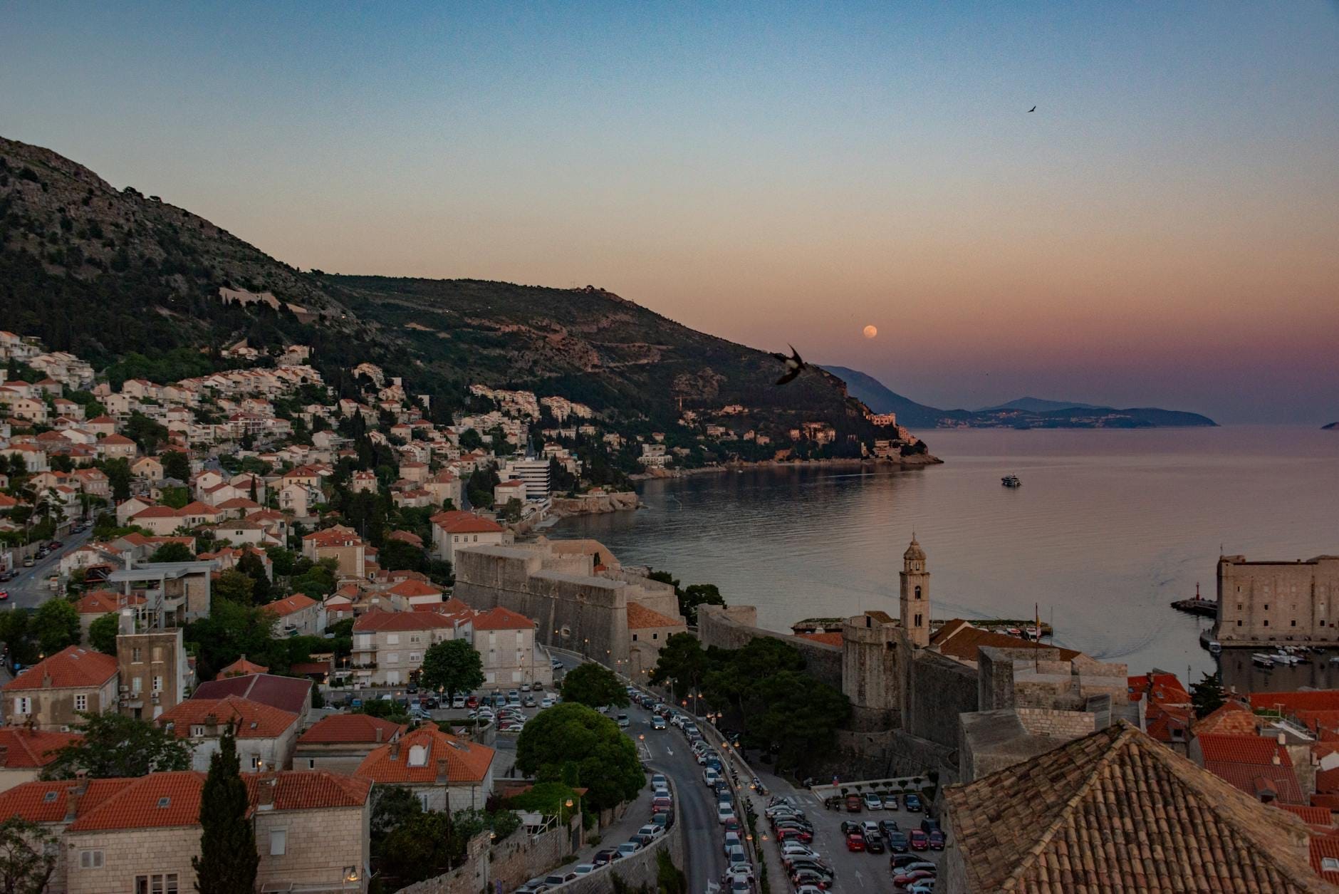 Vista elevada de Dubrovnik al anochecer con calles de la ciudad, murallas de la Ciudad Vieja, casas en la ladera y la costa del Adriático.