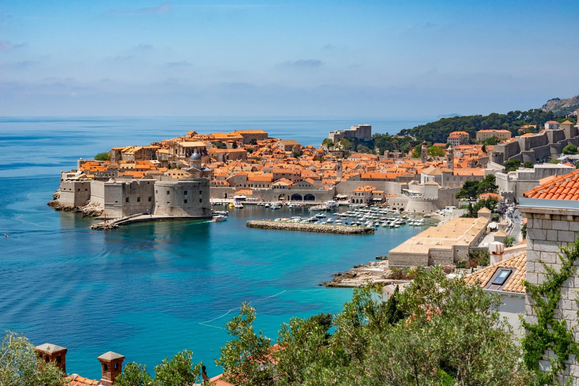 Vista panorámica impresionante de la Ciudad Vieja de Dubrovnik con sus icónicas murallas de piedra, tejados naranjas, el azul del mar Adriático, una pequeña marina y vegetación exuberante en primer plano.