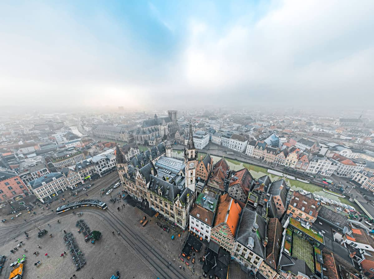 Wide aerial view of central Ghent showing historic buildings, multiple streets, tram tracks, and the rivers that define the old city layout under a partly cloudy sky.