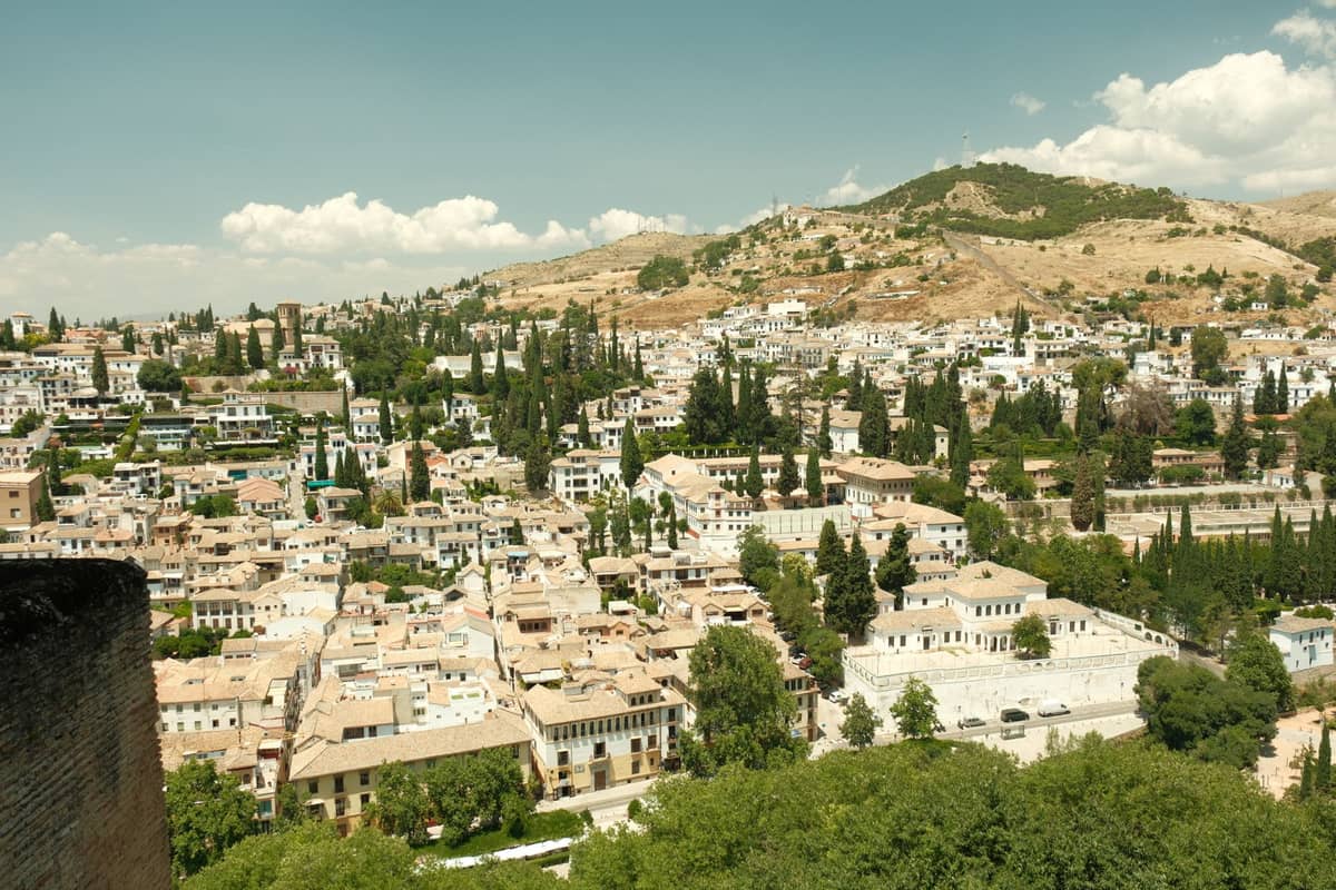 วิวพาโนรามาของเมือง Granada แสดงให้เห็นย่านที่อยู่อาศัยบนเนินเขาที่โดดเด่น พร้อมอาคารทาสีขาว ต้นไซเปรส และเนินเขาแห้งแล้งในพื้นหลังใต้ท้องฟ้าสีฟ้า