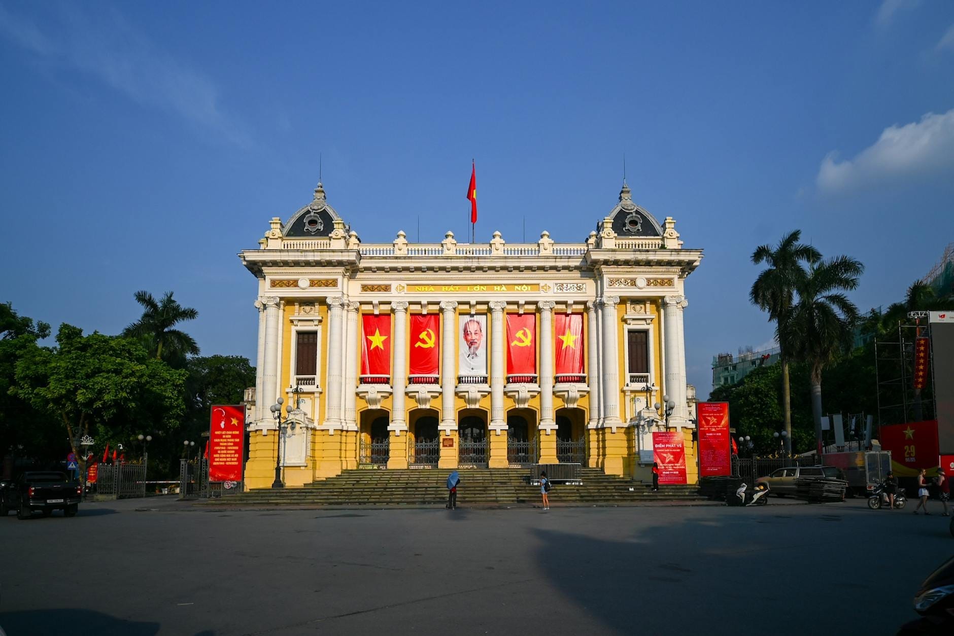 Front view of Hanoi Opera House with French colonial architecture, grand columns, and Vietnamese flags in a spacious, sunlit square.
