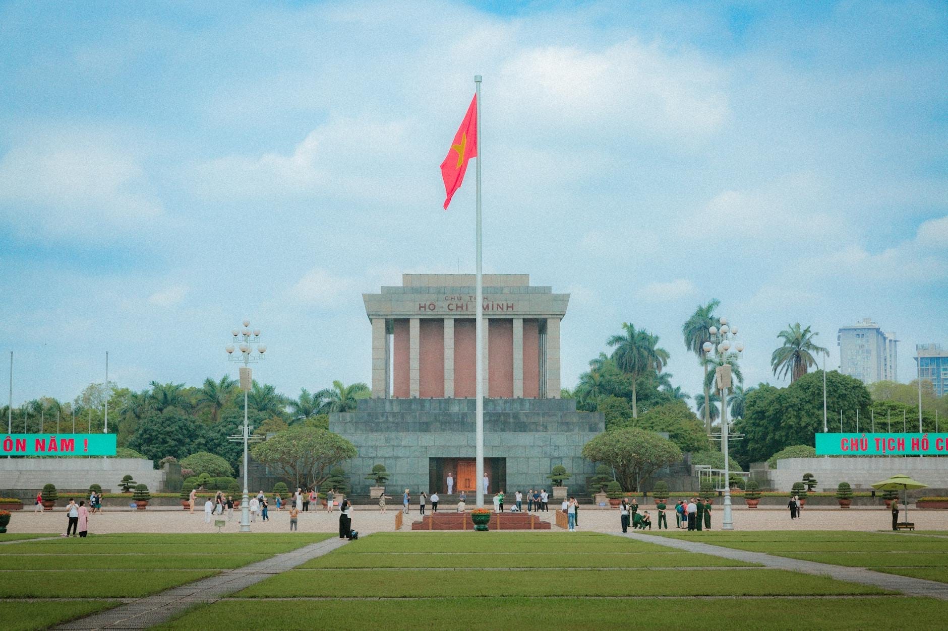 Wide daytime view of the Ho Chi Minh Mausoleum in Ba Dinh Square, Hanoi, with a Vietnamese flag, green lawns, and visitors walking nearby.